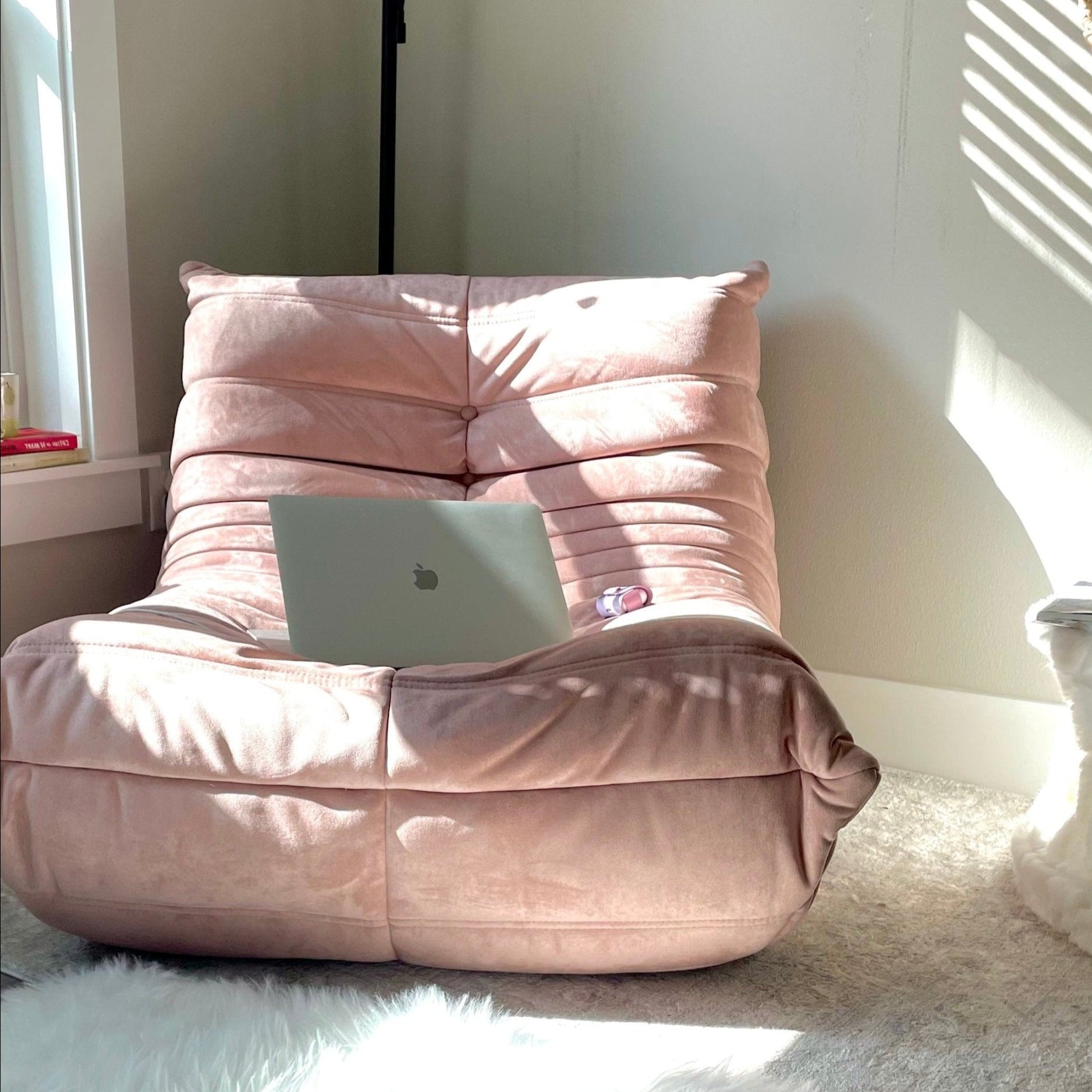 A pink Chillax Lounger Sofa Chair, featuring plush cushioning and holding a laptop, sits perfectly in a sunlit corner. The window blinds create playful patterns on the wall and floor, enhancing the luxurious lounging experience.