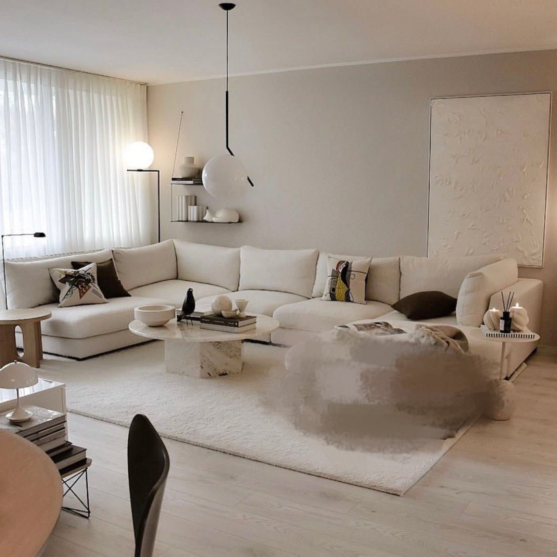 A modern living room showcases the Snowdrift L-Shaped Sectional Sofa in cream, abstract wall art, and a round marble coffee table on a light rug. Soft lighting and neutral tones create a cozy atmosphere.