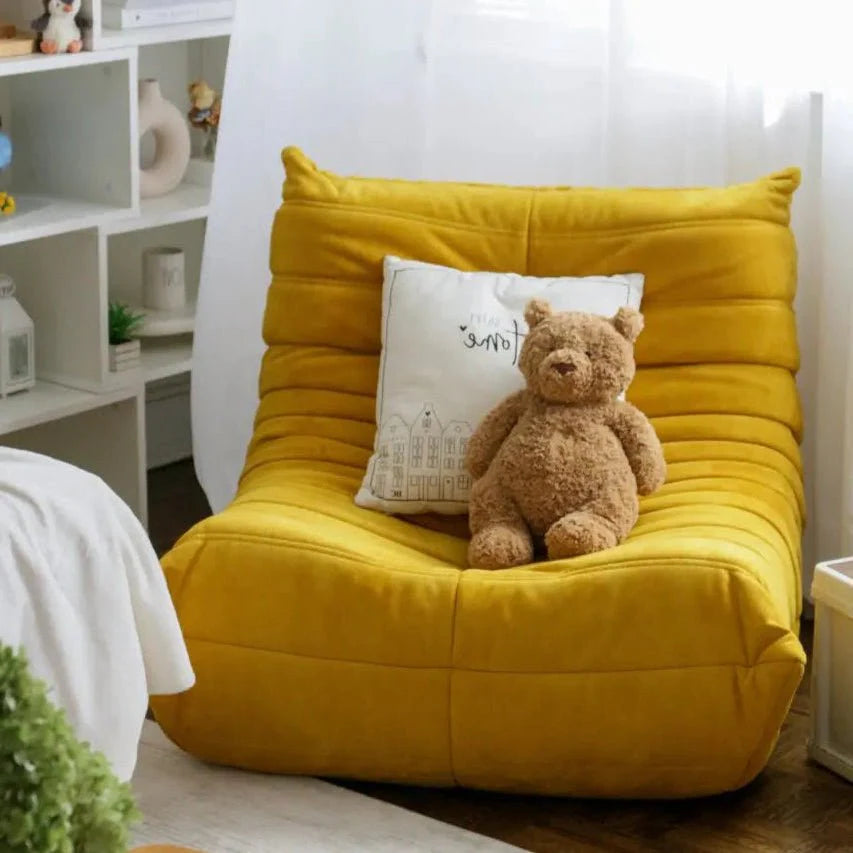 A plush teddy bear sits on a yellow Chillax Lounger Sofa Chair with ergonomic design and a decorative pillow. The room features a bookshelf adorned with decor, lit by natural sunlight.
