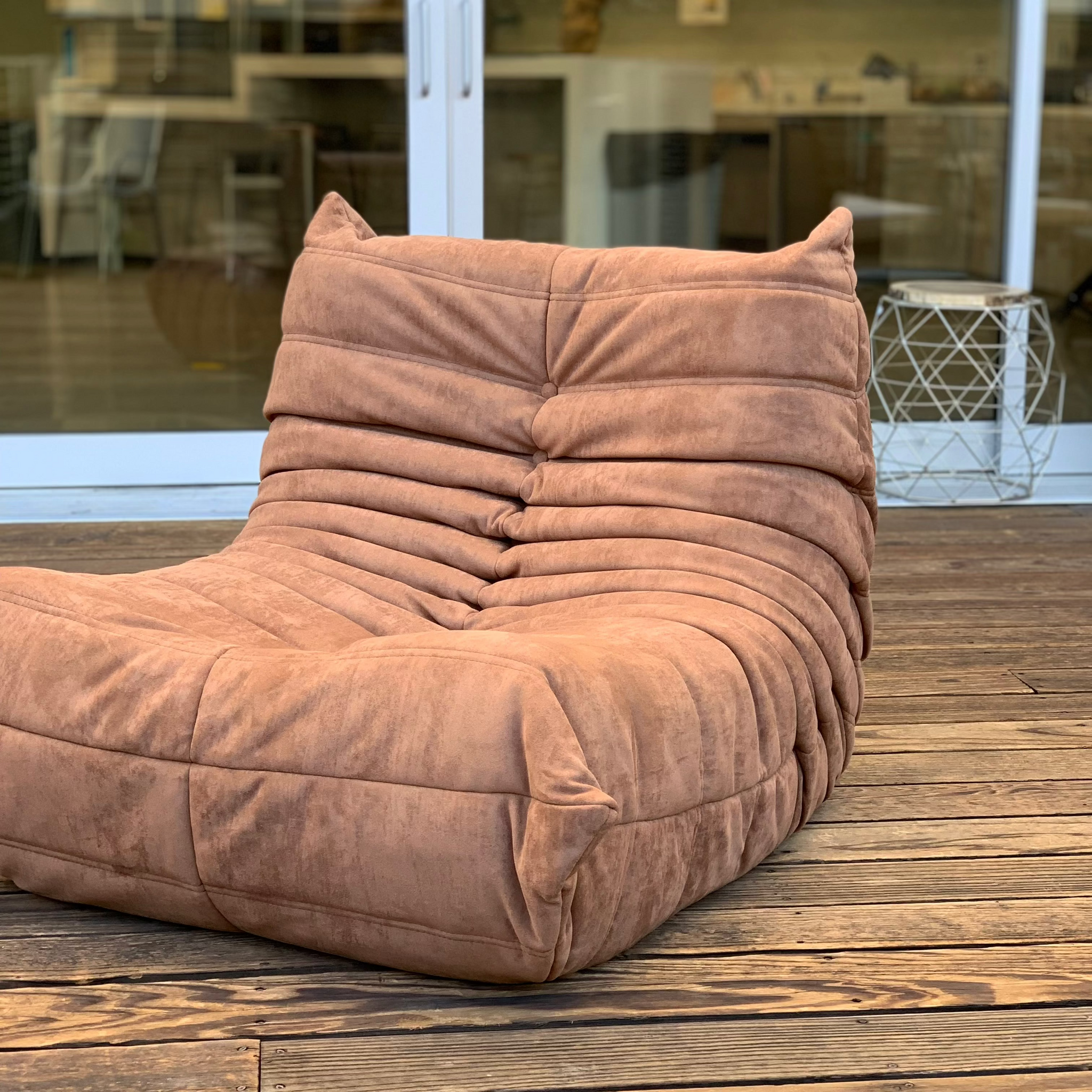 A cushioned Chillax Lounger Sofa Chair in brown rests on a wooden deck beside a glass door, with a metal side table in the background, providing a luxurious lounging experience.