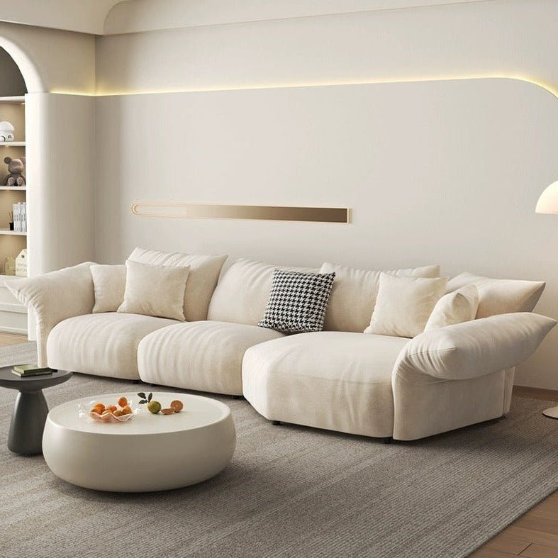 In the modern living room, a Petala Cream Sectional Sofa with Chaise takes center stage alongside a round white coffee table with oranges. A built-in wall shelf, ambient lighting, and a stylish floor lamp add warmth to the inviting space.