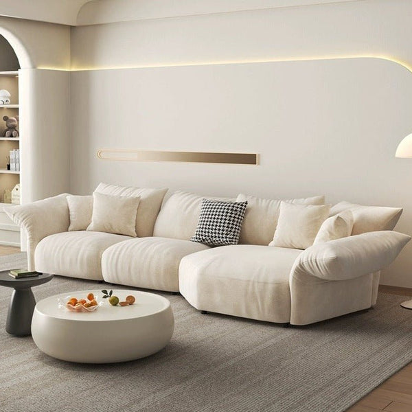 In the modern living room, a Petala Cream Sectional Sofa with Chaise takes center stage alongside a round white coffee table with oranges. A built-in wall shelf, ambient lighting, and a stylish floor lamp add warmth to the inviting space.
