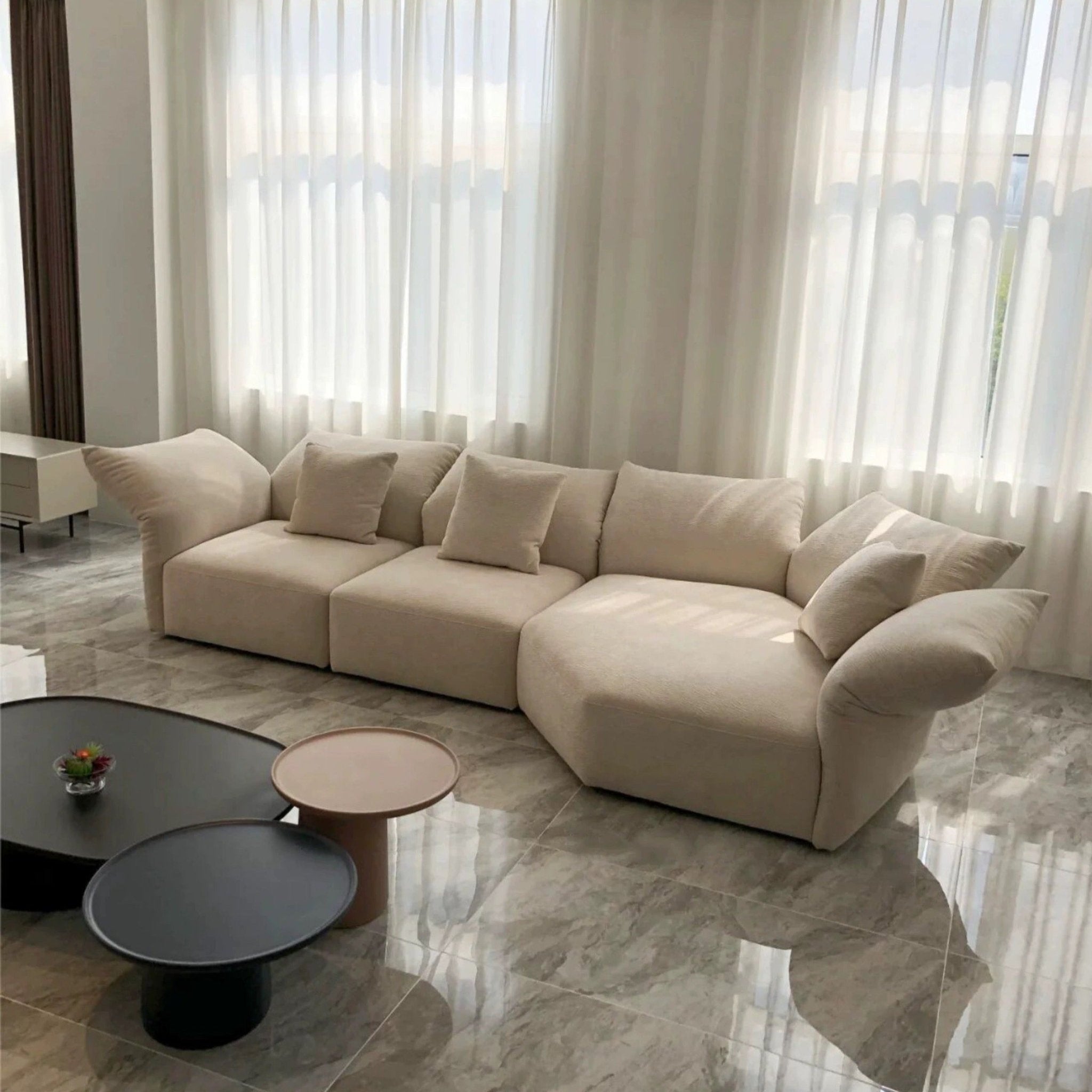 A contemporary silhouette enhances the bright room with a Petala Cream Modular Sofa with Chaise, adorned with cushions. In front, three round coffee tables of differing heights add to the scene, while white curtains drape elegantly over large windows.