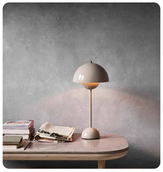 The Large Bud Table Lamp | Plug-In Edition with Cast Iron Base is illuminated on a wooden desk next to books, notebooks, and pens, set against a plain gray wall.