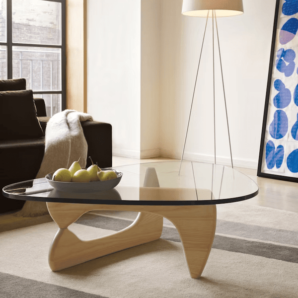 A modern living room features a Noguchi Coffee Table, a bowl of green apples, a floor lamp, a brown sofa with a blanket, and abstract wall art.