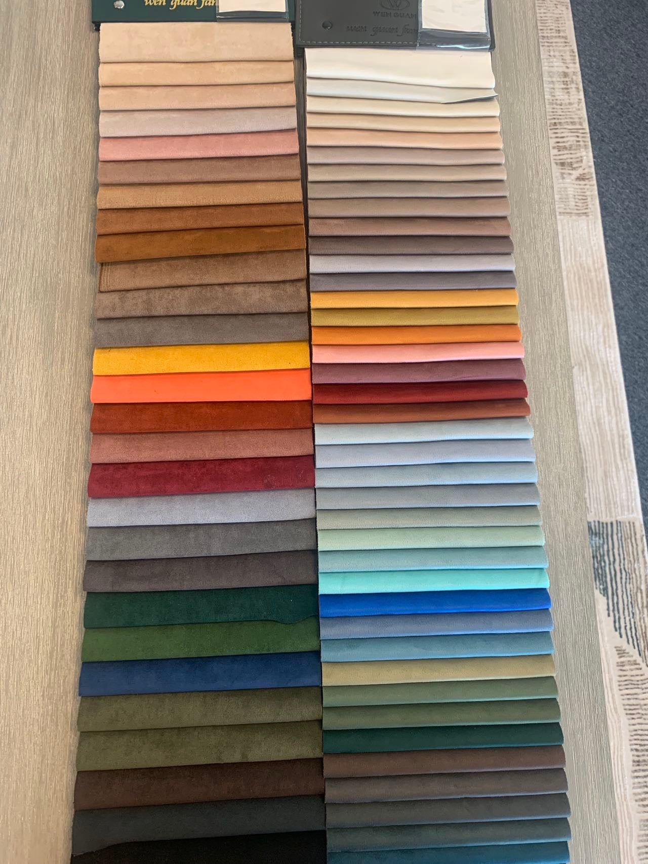 A table showcases neatly arranged fabric swatches in diverse colors, such as brown, red, orange, yellow, green, blue, and gray—perfectly complementing the luxurious experience on the Large Chillax Lounger – 2 Seat Sofa.
