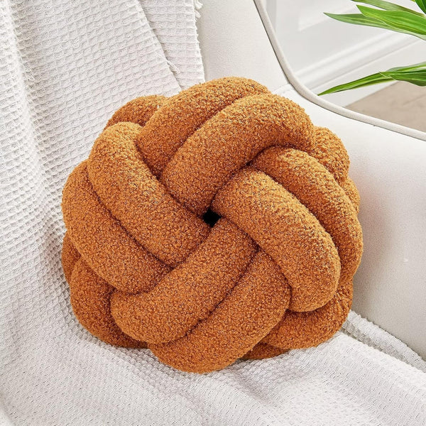The Knot Ball Pillow | Hand-Knotted Lamb Wool Throw Pillow in rust rests on a white textured blanket over a light chair beside a green plant, adding cozy modern style to the space.