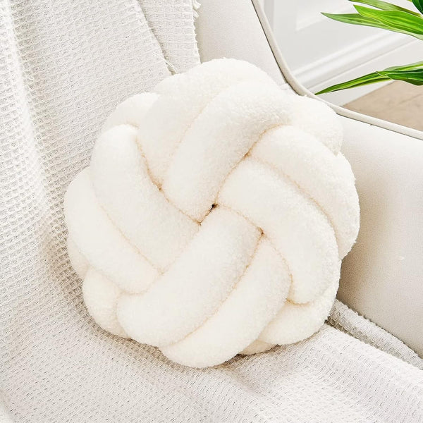The Knot Ball Pillow | Hand-Knotted Lamb Wool Throw Pillow for Cozy Modern Living sits on a textured blanket over a light chair, with a green plant partly visible in the background.